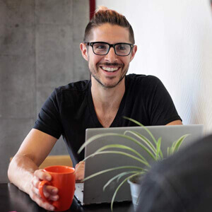 Smiling man in glasses sits at desk behind laptop