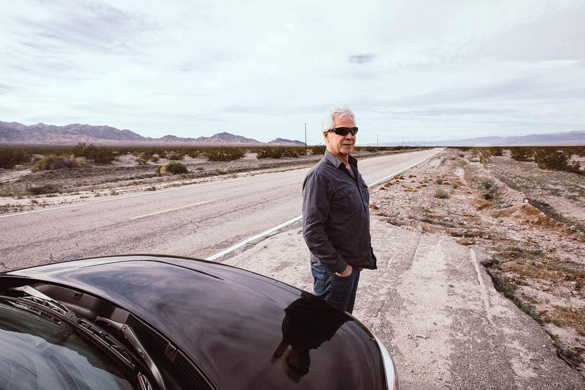 Older man wearing sunglasses stands before car admiring desert landscape