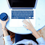 Top-down view of a woman using a MacBook while holding a cup of coffee